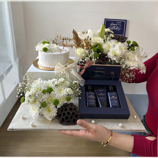 A gift hamper set including a round cake with white and green icing, surrounded by an assortment of fresh flowers such as white roses and daisies, and a box of bath care products which includes three small bottles.