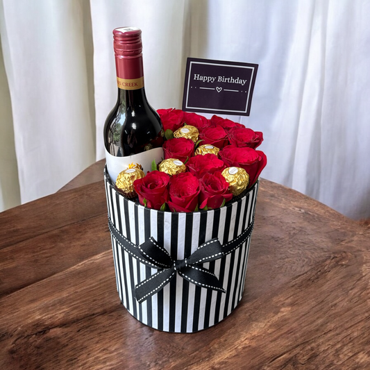Bottle of red wine with a bouquet of red roses and gold chocolate bars in a black and white striped box on a wooden surface.