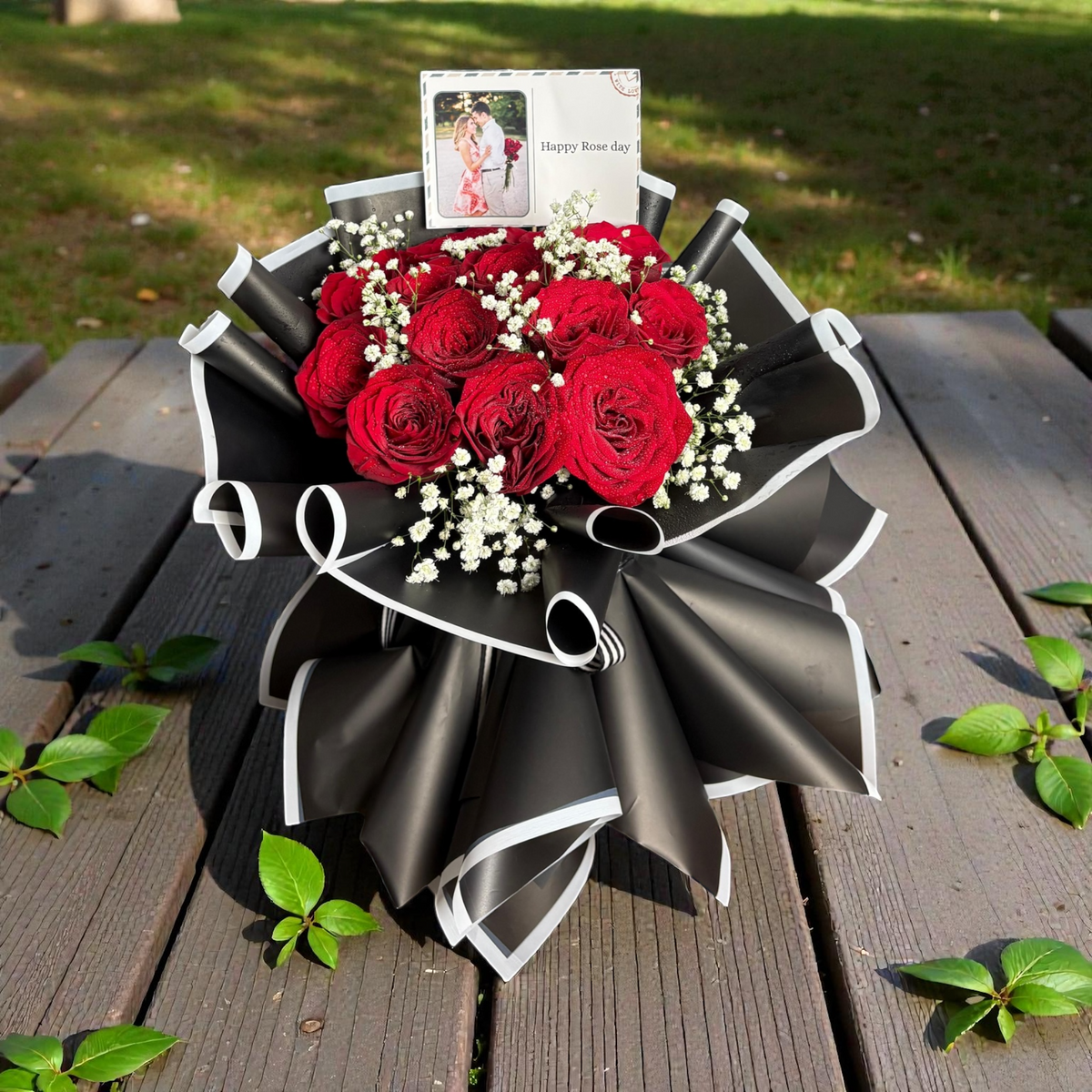 Bouquet of red roses wrapped in black paper on a wooden surface with greenery.