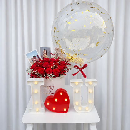 Decorative setup with red roses, balloons, and 'I love you' lights on a white surface.