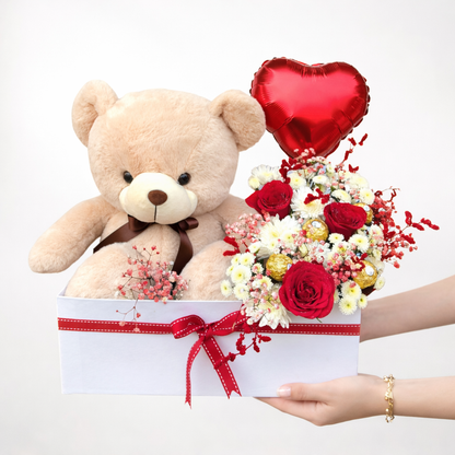 Teddy bear, bouquet of flowers, and heart-shaped balloon in a box held by hands on a white background