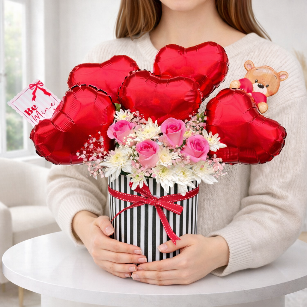 Person holding a bouquet of flowers with red heart-shaped balloons and a teddy bear in a box.