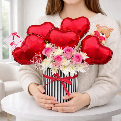 Person holding a bouquet of flowers with red heart-shaped balloons and a teddy bear in a box.