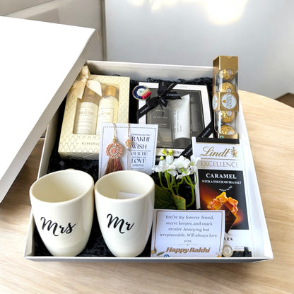 Gift box with 'Mr' and 'Mrs' mugs, Lindt chocolate, and other items on a wooden surface.