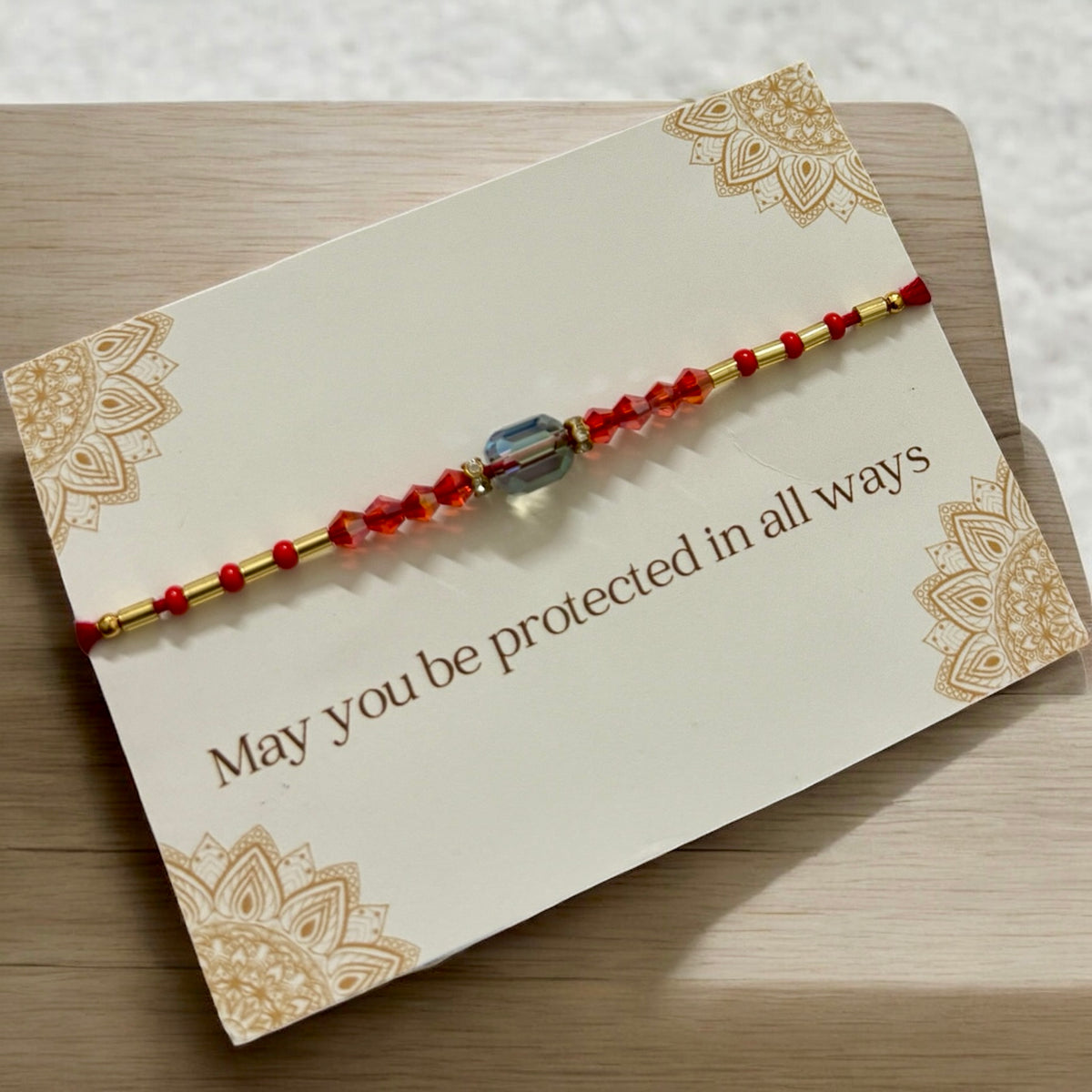 Bracelet with red beads and gold accents on a card with decorative patterns and text.