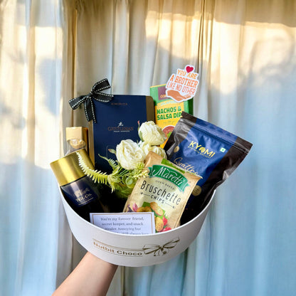Gourmet gift basket with snacks and flowers against a light curtain background