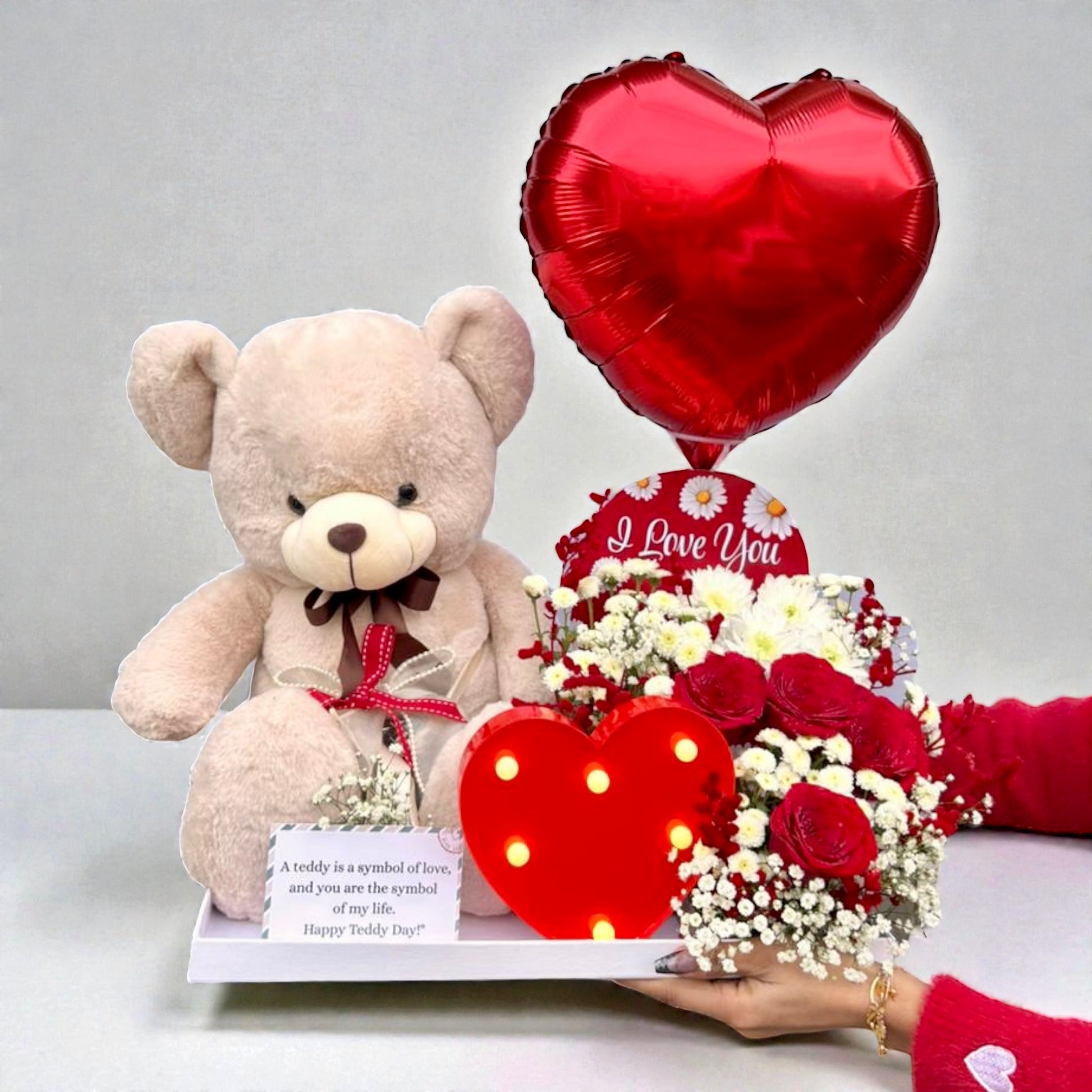 Teddy bear, bouquet of flowers, and heart-shaped balloon on a table with a gray background